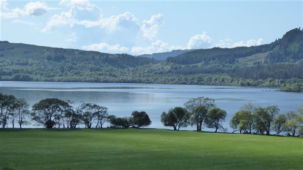 A green field with trees round the edge of the field and before a glistening loch and hills in the distance